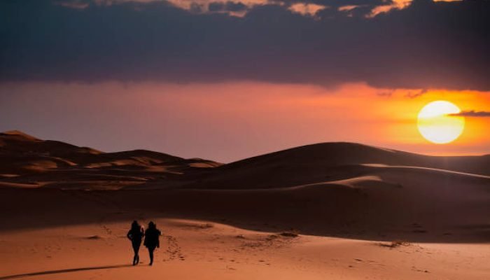 (Selective focus) Silhouette of two people walking on the sand dunes of the Merzouga desert during a stunning sunset. Merzouga, Morocco.